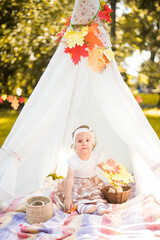 Fototapeta na wymiar Little girl lying and playing in a tent, children's house wigwam in park Autumn portrait of cute curly girl. Harvest or Thanksgiving. autumn decor, party, picnic. Child in white dress with pumpkins. 