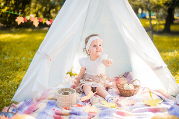 Little girl lying and playing in a tent, children's house wigwam in park Autumn portrait of cute curly girl. Harvest or Thanksgiving. autumn decor, party, picnic. Child in white dress with pumpkins.  © MartaKlos