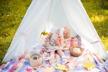 Little girl lying and playing in a tent, children's house wigwam in park Autumn portrait of cute curly girl. Harvest or Thanksgiving. autumn decor, party, picnic. Child in white dress with pumpkins.  © MartaKlos