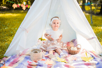 Little girl lying and playing in a tent, children's house wigwam in park Autumn portrait of cute curly girl. Harvest or Thanksgiving. autumn decor, party, picnic. Child in white dress with pumpkins.  © MartaKlos