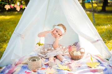 Little girl lying and playing in a tent, children's house wigwam in park Autumn portrait of cute curly girl. Harvest or Thanksgiving. autumn decor, party, picnic. Child in white dress with pumpkins.  © MartaKlos