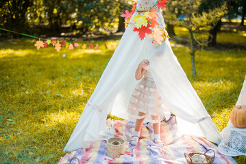 Little girl lying and playing in a tent, children's house wigwam in park Autumn portrait of cute curly girl. Harvest or Thanksgiving. autumn decor, party, picnic. Child in white dress with pumpkins.  © MartaKlos