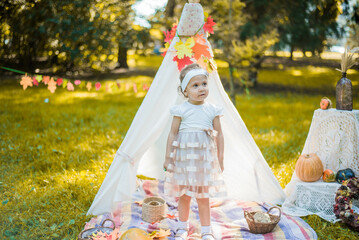 Little girl lying and playing in a tent, children's house wigwam in park Autumn portrait of cute curly girl. Harvest or Thanksgiving. autumn decor, party, picnic. Child in white dress with pumpkins.  © MartaKlos