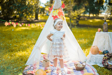 Little girl lying and playing in a tent, children's house wigwam in park Autumn portrait of cute curly girl. Harvest or Thanksgiving. autumn decor, party, picnic. Child in white dress with pumpkins.  © MartaKlos