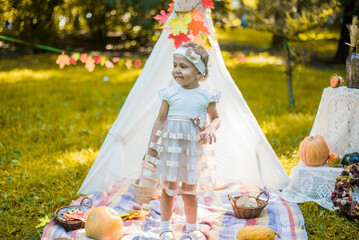Little girl lying and playing in a tent, children's house wigwam in park Autumn portrait of cute curly girl. Harvest or Thanksgiving. autumn decor, party, picnic. Child in white dress with pumpkins.  © MartaKlos