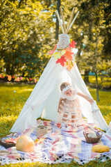 Little girl lying and playing in a tent, children's house wigwam in park Autumn portrait of cute curly girl. Harvest or Thanksgiving. autumn decor, party, picnic. Child in white dress with pumpkins.  © MartaKlos