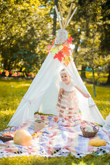 Little girl lying and playing in a tent, children's house wigwam in park Autumn portrait of cute curly girl. Harvest or Thanksgiving. autumn decor, party, picnic. Child in white dress with pumpkins.  © MartaKlos
