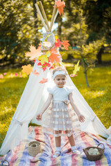 Little girl lying and playing in a tent, children's house wigwam in park Autumn portrait of cute curly girl. Harvest or Thanksgiving. autumn decor, party, picnic. Child in white dress with pumpkins.  © MartaKlos