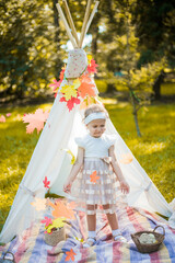 Little girl lying and playing in a tent, children's house wigwam in park Autumn portrait of cute curly girl. Harvest or Thanksgiving. autumn decor, party, picnic. Child in white dress with pumpkins.  © MartaKlos