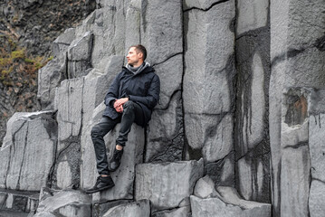 Male tourist looking away while resting on beautiful basalt columns. Scenic view of gray patterned stones at famous Reynisfjara Beach. Explorer spending leisure time at rocky cliff.