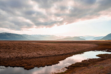 View of stream flowing amidst grassy landscape. Beautiful view of mountain ranges in volcanic valley against cloudy sky. Beautiful scenery of dramatic field during sunset.