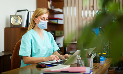 Professional general practitioner woman in mask working sitting at working place in office behind laptop