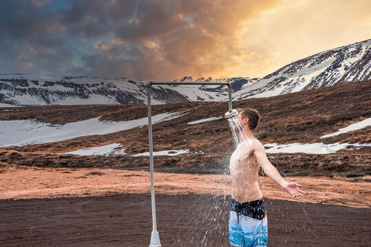 Young tourist showering in hot shower station from geothermal power at Krafla. Shirtless traveler enjoying bath against snow covered mountain. He is spending leisure time against sky during sunset.