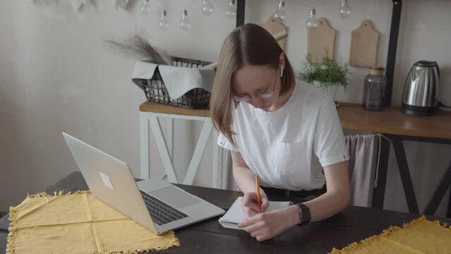 Close Up Serious Focused Woman With Ear Buds Looking At Laptop Screen, Sitting At Desk, Writing In Notepad. 