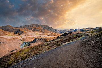 Scenic view of hot streams amidst landscape against cloudy sky. Distant flowing water from mountain in Hveragerdi valley. Idyllic scenery of volcanic land during sunset.