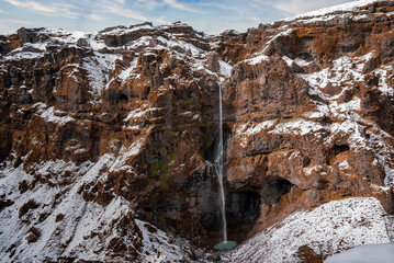 Beautiful small waterfall falling from snow covered mountains. Majestic rocky cliffs against sky. Idyllic view of Icelandic nature in volcanic valley.