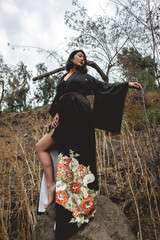 Portrait of sexy and young japanese woman with beautiful old traditional black kimono with flowers posing and showing legs and feet over a big rock in the autumn forest 