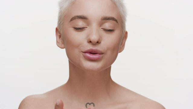Gorgeous young slim light-haired Caucasian woman with naked shoulders blows a kiss for the camera on white background | Lips care concept