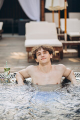 Curly positive guy holds cocktails and enjoys spending time in the jacuzzi