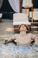 Curly positive guy holds cocktails and enjoys spending time in the jacuzzi