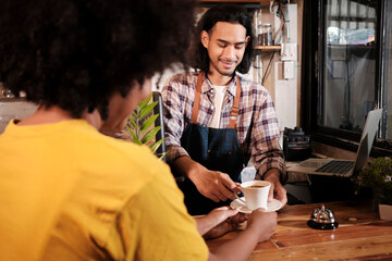 Close-up shot, young long hair Thai male baristas startup works by serving a cup of coffee to a customer with a smile at casual cafe counter bar, happy service job, and small business entrepreneur.