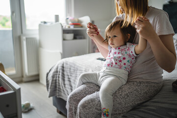 Mother and daughter caucasian woman hold her small toddler while sitting on the bed at home in...