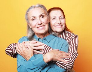 Two pretty older women hugging on yellow background