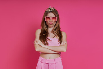 Angry young woman in funky crown frowning and looking angry while standing against pink background