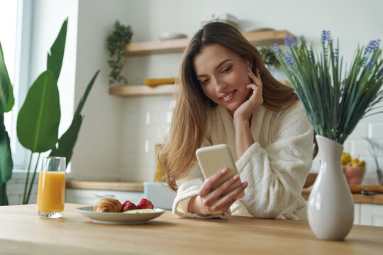 Beautiful young woman in bathrobe using smart phone while spending time at the modern kitchen