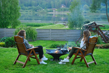Two unrecognizable women in the backyard, sitting in chairs and chatting near the barbecue