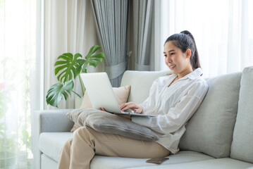 Image of cheerful asian woman using laptop while sitting on couch in living room. She uses her laptop for meetings, searching for information, chatting with friends, and enjoying shopping online.