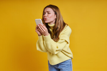 Beautiful woman holding smart phone and sending a kiss while standing against yellow background