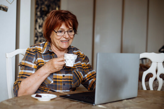 Smiling senior woman in glasses, using laptop at home. Drinking tea or coffee. Working at home, computer technology. 