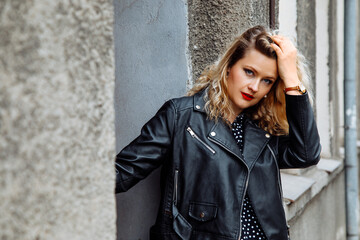 Obraz premium Modern stylish blond woman with make-up posing in cool black leather jacket and dress against wall. Confrontation, rebel