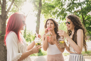 Three young women in fashionable clothes and bright sunglasses walk around the city in summer and drink refreshing non-alcoholic cocktails from disposable plastic cups. Women's friendship and walks in