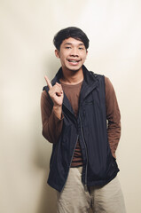 Portrait of Asian young man pointing at empty space wearing brown t-shirt and black vest isolated on background