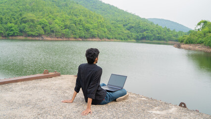 Young man working on a laptop and sitting in the nature, Workation and Digital Nomad concept.