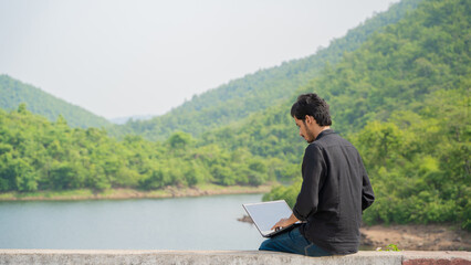 Fototapeta premium Young man working on a laptop and sitting in the nature, Workation and Digital Nomad concept.