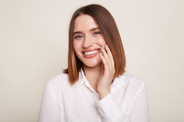 Portrait of smiling cheerful Caucasian woman wearing shirt posing isolated over white background, looking at camera with happy facial expression, keeps hand on cheek.