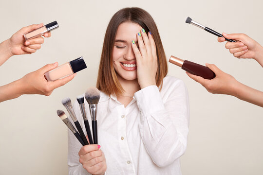 Image of the hands of several beauticians holding their respective equipment giving makeover to beautiful smiling woman with charming smile, standing and covering eye with palm.