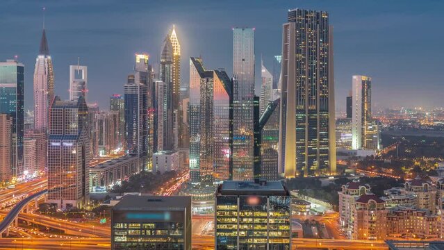 Panorama of Dubai Financial Center district with tall skyscrapers with illumination day to night transition timelapse. Aerial view to towers along busy highway after sunset