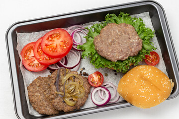 Fried burger bun on lettuce leaf, fried burger with cheese and fried onions, chopped tomatoes and onions on baking tray