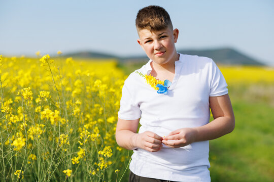 Teenage guy in T-shirt with Ukrainian flowers on it enjoys sun while standing in rapeseed field against clear sky