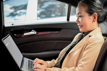 Successful aged asian businesswoman using portable laptop, working in the car during a rush hour