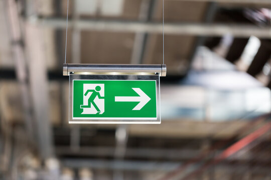 Emergency exit sign. With running figure and arrow to the right. Blurry background, hanging from the ceiling.