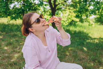 Funny brunette girl in sunglasses eating pizza on nature background. Attractive girl feels hungry and loves the taste. Entire pizza for myself. Fast food banner