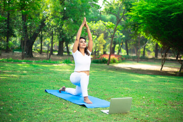 Young indian woman doing fitness stretching yoga exercise in summer park while using laptop for...