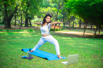 Young indian woman doing fitness stretching yoga exercise in the park while using laptop for online class or virtual tutorials. healthy lifestyle.