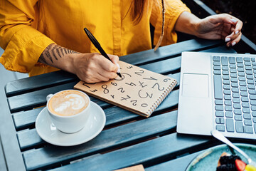 Woman freelancer writing numbers in notepad while working with laptop in street cafe. Time Management