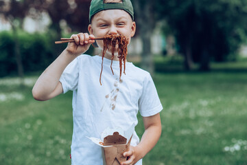 Boy holding noodles rolled up on chopsticks closes his mouth, outdoors. Dirty stain of sauce on his...